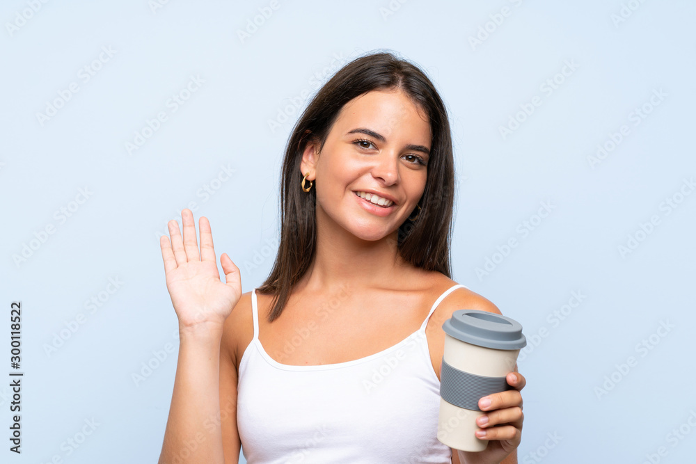 Young woman holding a take away coffee over isolated blue background saluting with hand with happy expression Young woman holding a take away coffee over isolated blue background saluting with hand with happy expression