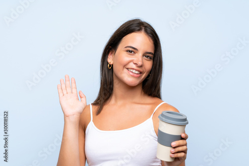 Young woman holding a take away coffee over isolated blue background saluting with hand with happy expression