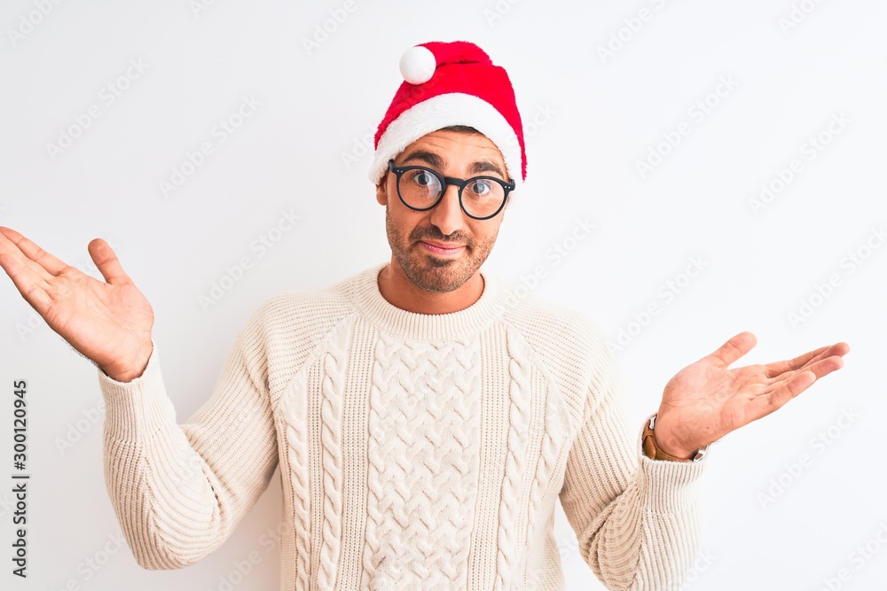 Naklejka premium Young handsome man wearing christmas hat and glasses over isolated background clueless and confused expression with arms and hands raised. Doubt concept.