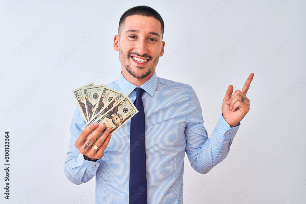 Young handsome business man holding a bunch of bank notes dollars over isolated background very happy pointing with hand and finger to the side Young handsome business man holding a bunch of bank notes dollars over isolated background very happy pointing with hand and finger to the side