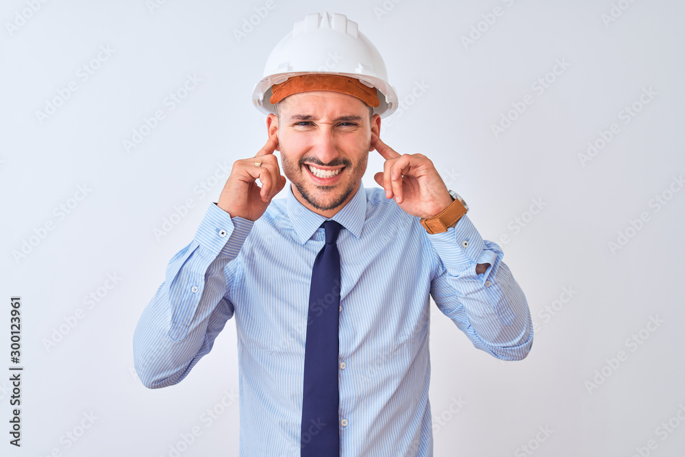 Young business man wearing contractor safety helmet over isolated background covering ears with fingers with annoyed expression for the noise of loud music. Deaf concept. Young business man wearing contractor safety helmet over isolated background covering ears with fingers with annoyed expression for the noise of loud music. Deaf concept.