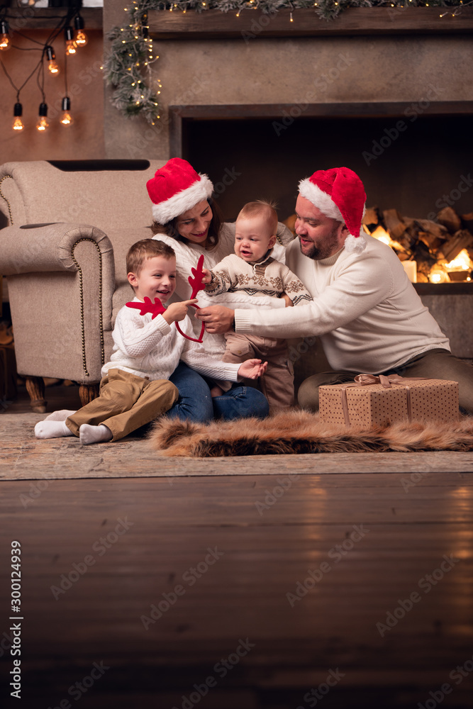 Obraz premium Image of parents in Santa's cap with two sons sitting on floor by fireplace in room