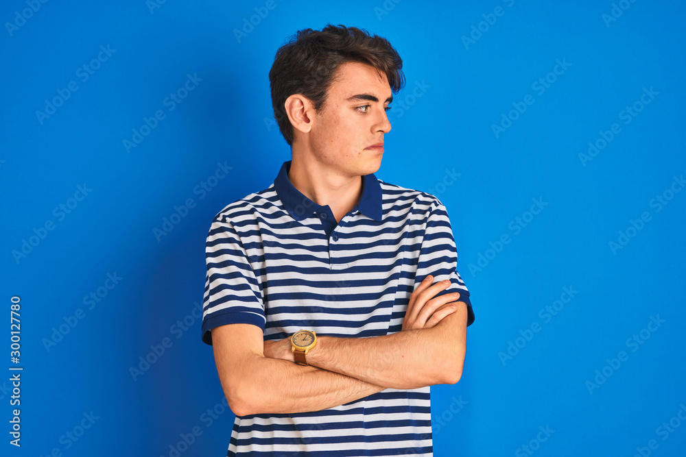 Teenager boy wearing casual t-shirt standing over blue isolated background looking to the side with arms crossed convinced and confident Teenager boy wearing casual t-shirt standing over blue isolated background looking to the side with arms crossed convinced and confident