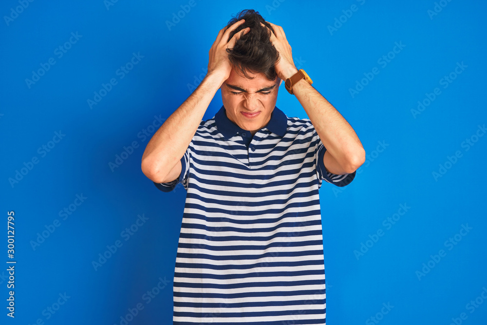 Teenager boy wearing casual t-shirt standing over blue isolated background suffering from headache desperate and stressed because pain and migraine. Hands on head. Teenager boy wearing casual t-shirt standing over blue isolated background suffering from headache desperate and stressed because pain and migraine. Hands on head.