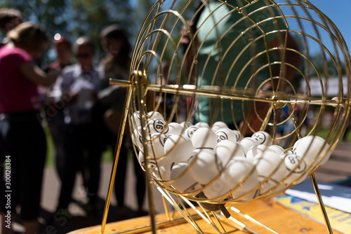 Bingo balls on a table next to a lotto raffle drum machine. Group on people playing lottery in outdoor backyard setup at an event or party.