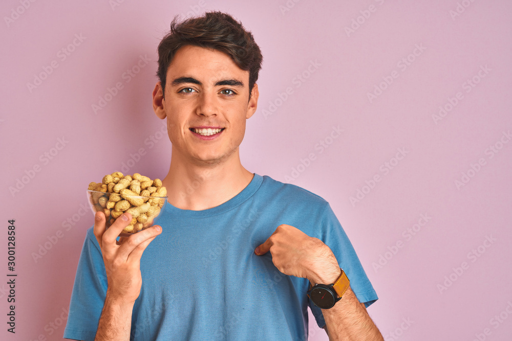 Teenager boy holding a bunch of peanunts over isolated pink background with surprise face pointing finger to himself Teenager boy holding a bunch of peanunts over isolated pink background with surprise face pointing finger to himself