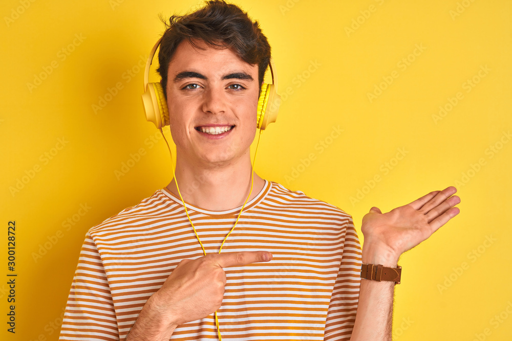 Teenager boy wearing headphones over isolated yellow background amazed and smiling to the camera while presenting with hand and pointing with finger. Teenager boy wearing headphones over isolated yellow background amazed and smiling to the camera while presenting with hand and pointing with finger.