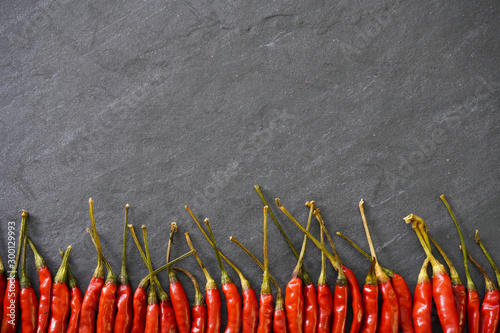 A row of bright red Thai bird chiles rest on black slate to be prepared for a meal of bright red Thai bird chiles with green stems against a dark gray slate background with copy space