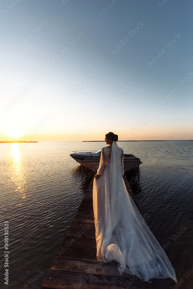 Beautiful bride in an elegant white dress on a bridge against the background of the sea and in the rays of sunset. Happy bride with luxury make-up and hairstyle. Wedding day.