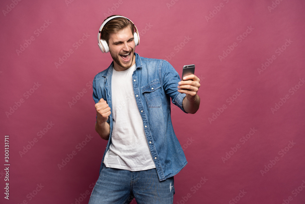 Excited young man standing isolated over pink background, listening to music with earphones and mobile phone Excited young man standing isolated over pink background, listening to music with earphones and mobile phone