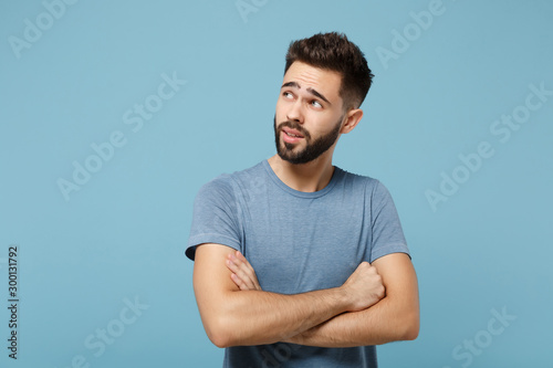 Young pensive handsome man in casual clothes posing isolated on blue background, studio portrait. People sincere emotions lifestyle concept. Mock up copy space. Holding hands crossed, looking aside.