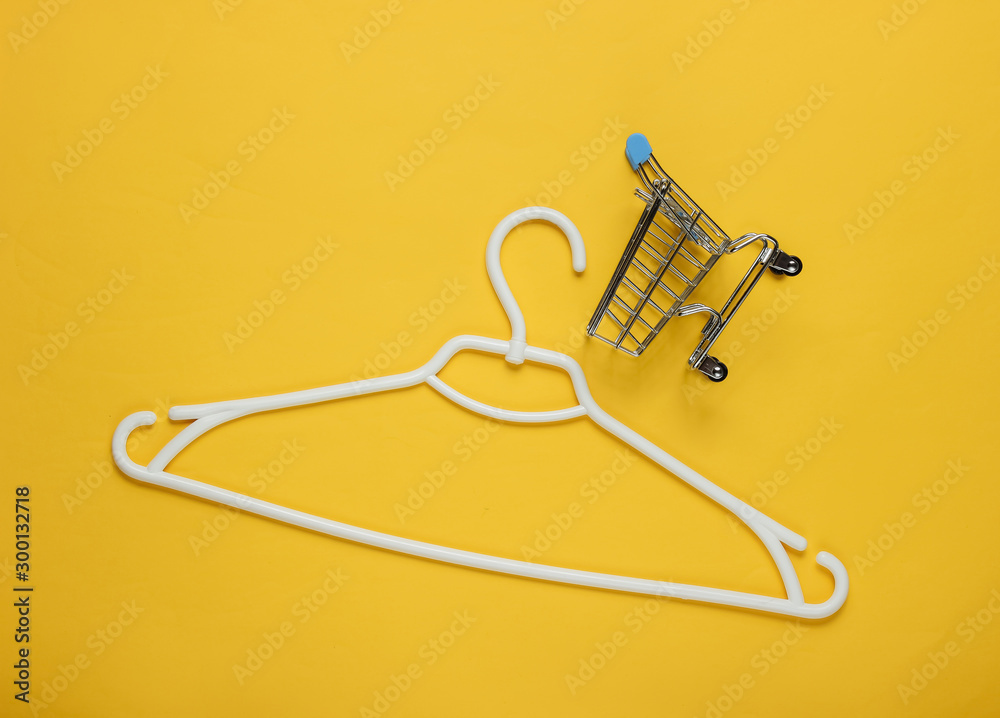 Fototapeta premium Plastic hanger and shopping trolley on yellow paper background. Minimalistic fashion concept. Top view