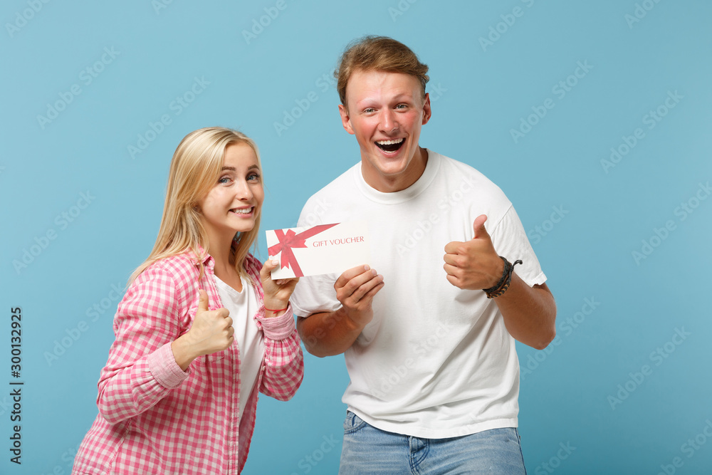 Young laughing couple two friends guy girl in white pink empty t-shirts posing isolated on pastel blue background. People lifestyle concept. Mock up copy space. Hold gift certificate showing thumb up. Young laughing couple two friends guy girl in white pink empty t-shirts posing isolated on pastel blue background. People lifestyle concept. Mock up copy space. Hold gift certificate showing thumb up.
