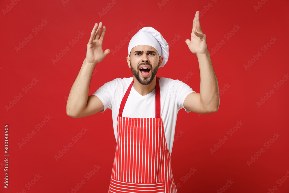 Irritated young bearded male chef cook or baker man in striped apron toque chefs hat posing isolated on red background. Cooking food concept. Mock up copy space. Screaming, swearing, spreading hands. Irritated young bearded male chef cook or baker man in striped apron toque chefs hat posing isolated on red background. Cooking food concept. Mock up copy space. Screaming, swearing, spreading hands.