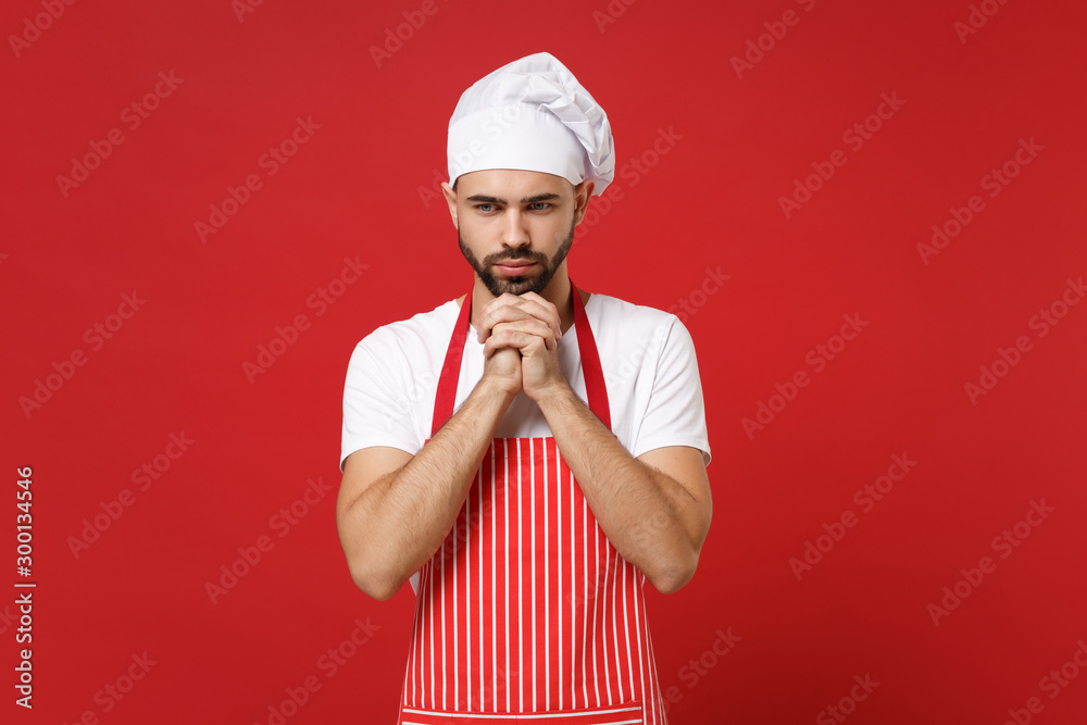Pensive male chef cook baker man in striped apron white t-shirt toque chefs hat posing isolated on red background. Cooking food concept. Mock up copy space. Put hands prop up on chin, looking down. Pensive male chef cook baker man in striped apron white t-shirt toque chefs hat posing isolated on red background. Cooking food concept. Mock up copy space. Put hands prop up on chin, looking down.