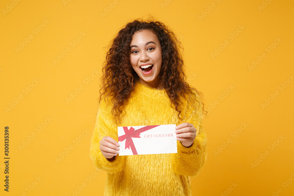 Excited young african american girl in fur sweater posing isolated on yellow orange background studio portrait. People lifestyle concept. Mock up copy space. Hold gift certificate, keeping mouth open. - obrazy, fototapety, plakaty 