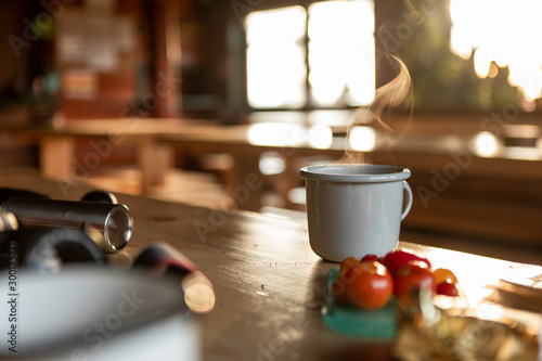 Grey enamel cup of hot steaming coffee or tea sitting on an old table in a mountain shelter. © Lukasz