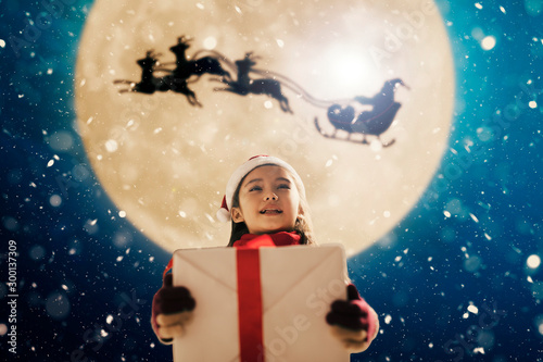 Cute little girl with christmas presents. Santa Claus flying in  moon sky