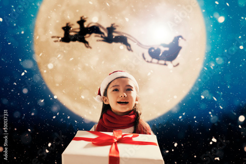 Cute little girl with christmas presents. Santa Claus flying in moon sky