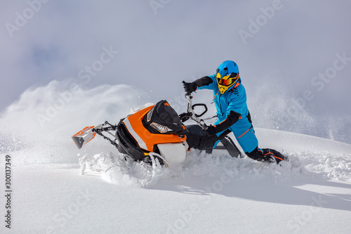 the guy turns a snowmobile in a mountain valley on the background of the clear snow and sky, leaving behind a trail of splashes. bright snow bike and suit without brands. Boondocker sports snowmobile © Wlad Go