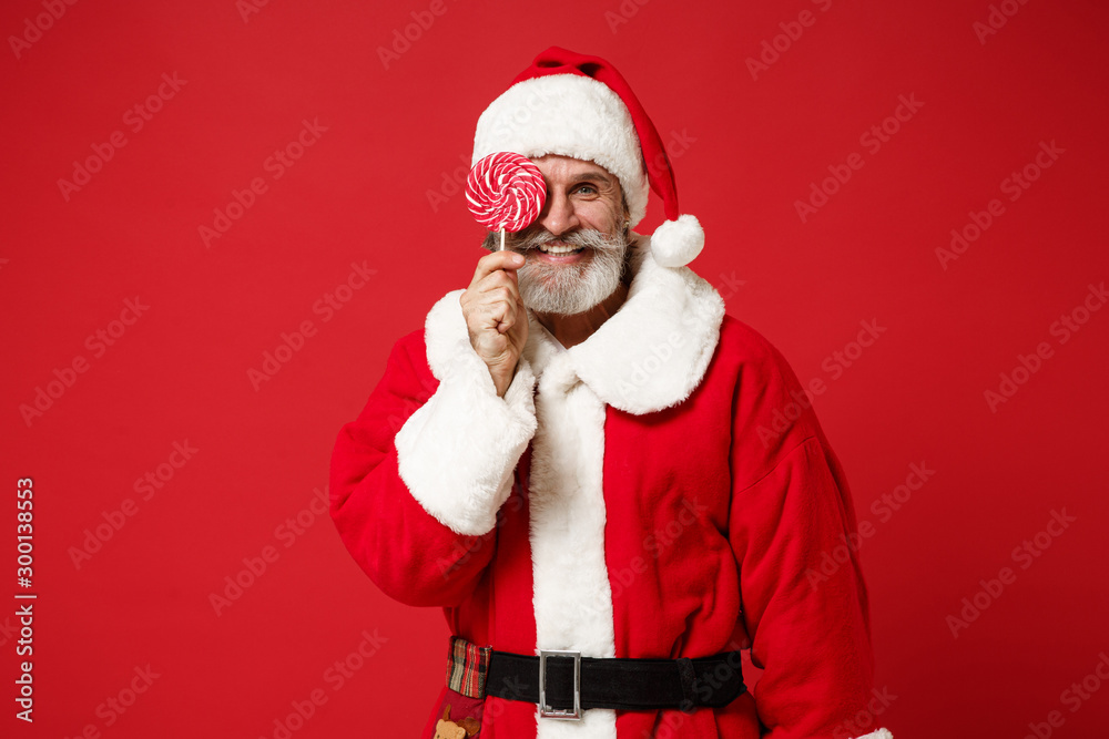Elderly gray-haired mustache bearded Santa man in Christmas hat posing isolated on red wall background. New Year 2020 celebration holiday concept. Mock up copy space. Covering eye with round lollipop. Elderly gray-haired mustache bearded Santa man in Christmas hat posing isolated on red wall background. New Year 2020 celebration holiday concept. Mock up copy space. Covering eye with round lollipop.