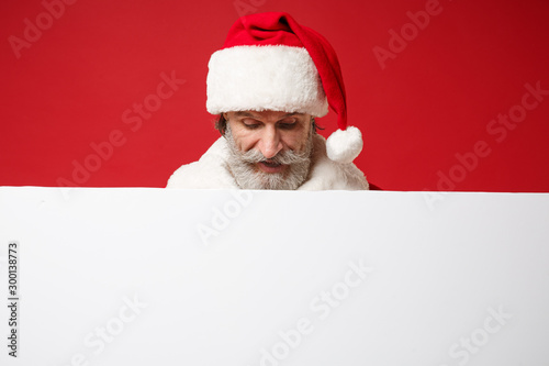 Elderly gray-haired mustache bearded Santa man in Christmas hat posing isolated on red background. Happy New Year 2020 celebration holiday concept. Mock up copy space. Holding white blank billboard. © ViDi Studio