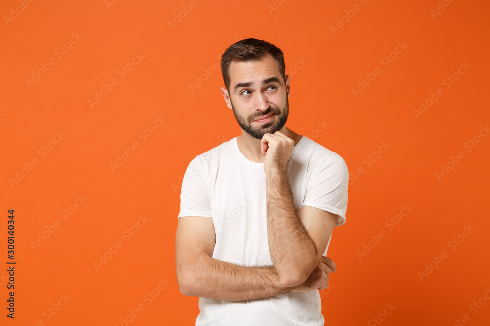Confused pensive young man in casual white t-shirt posing isolated on orange background, studio portrait. People sincere emotions lifestyle concept. Mock up copy space. Putting hand prop up on chin. Confused pensive young man in casual white t-shirt posing isolated on orange background, studio portrait. People sincere emotions lifestyle concept. Mock up copy space. Putting hand prop up on chin.