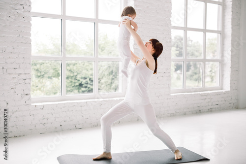 Sport, motherhood and active lifestyle concept - side view of young mother workout together with her baby over white wall and big windows background. Mother having fun and playing with her baby.