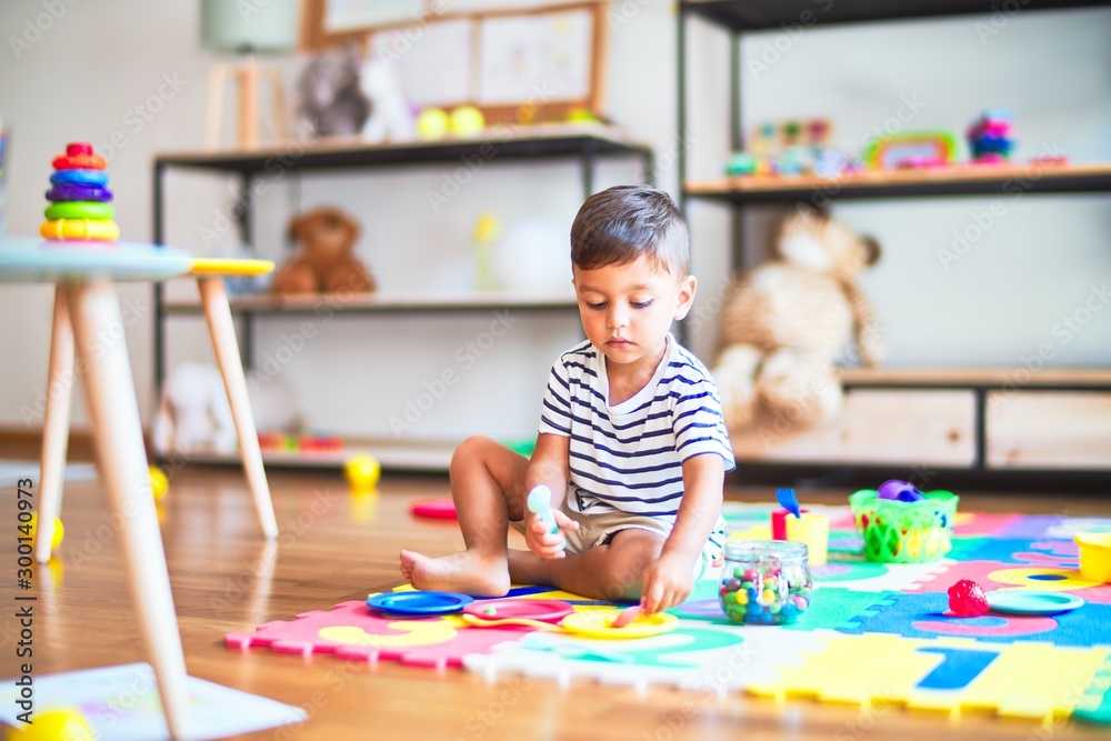 Fototapeta premium Beautiful toddler boy sitting on puzzle playing meals with plastic plates, fruits and vegetables at kindergarten