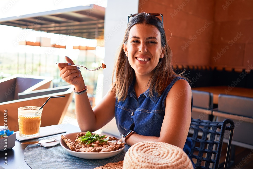 Young beautiful woman sitting at restaurant enjoying summer vacation eating delicious food - obrazy, fototapety, plakaty 