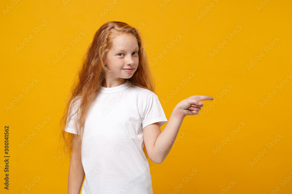 Smiling little ginger kid girl 12-13 years old in white t-shirt isolated on yellow background children studio portrait. Childhood lifestyle concept. Mock up copy space. Pointing index finger aside. Smiling little ginger kid girl 12-13 years old in white t-shirt isolated on yellow background children studio portrait. Childhood lifestyle concept. Mock up copy space. Pointing index finger aside.