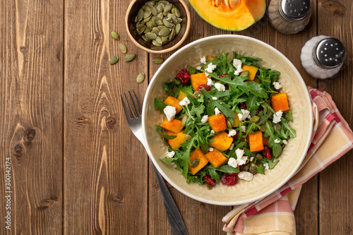 Autumn salad with baked pumpkin, arugula, seeds, dried cranberries and feta cheese in bowl on rustic wooden background. Top view. Copy space.