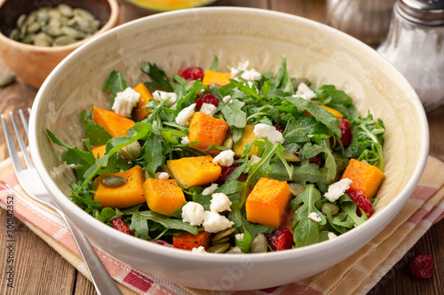 Autumn salad with baked pumpkin, arugula, seeds, dried cranberries and feta cheese in bowl on rustic wooden background. Selective focus.