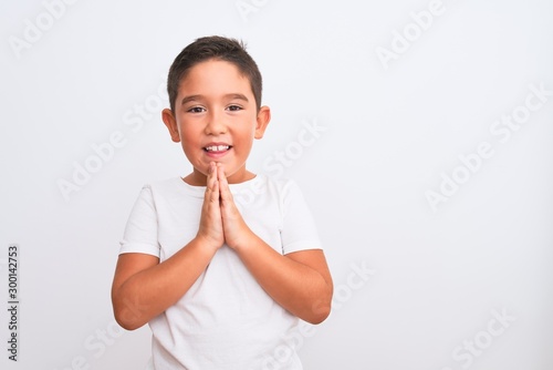 Beautiful kid boy wearing casual t-shirt standing over isolated white background praying with hands together asking for forgiveness smiling confident.