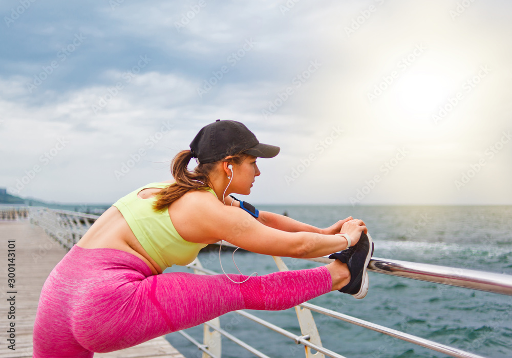 Sports woman in sportswear with headphones in the ears doing stretching leg  exercise before training on the beach outdoor