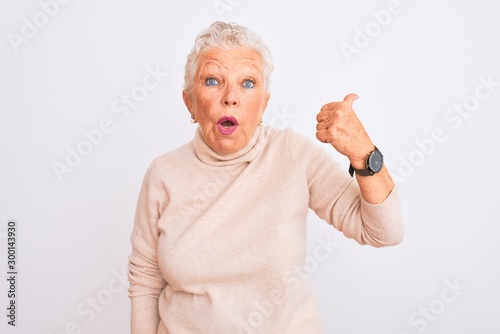 Senior grey-haired woman wearing turtleneck sweater standing over isolated white background Surprised pointing with hand finger to the side, open mouth amazed expression.