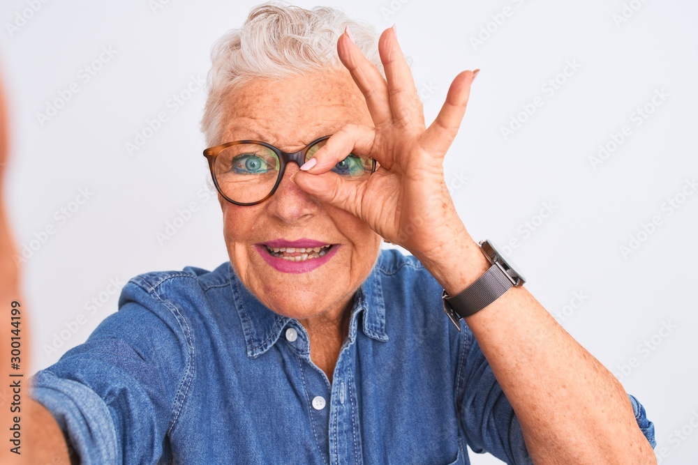 Grey-haired woman wearing denim shirt and glasses make selfie over isolated white background with happy face smiling doing ok sign with hand on eye looking through fingers Grey-haired woman wearing denim shirt and glasses make selfie over isolated white background with happy face smiling doing ok sign with hand on eye looking through fingers