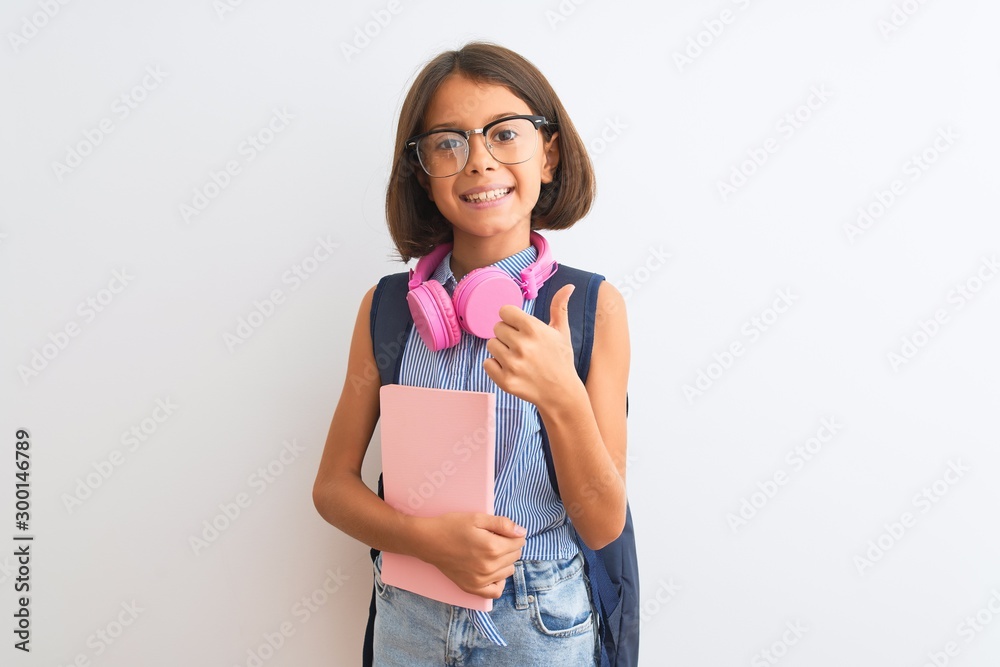 Student child girl wearing backpack glasses book headphones over isolated white background happy with big smile doing ok sign, thumb up with fingers, excellent sign Student child girl wearing backpack glasses book headphones over isolated white background happy with big smile doing ok sign, thumb up with fingers, excellent sign