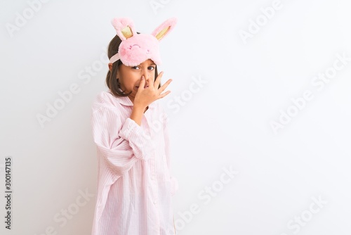Beautiful child girl wearing sleep mask and pajama standing over isolated white background smelling something stinky and disgusting, intolerable smell, holding breath with fingers on nose. Bad smell