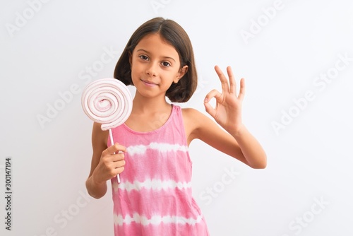 Beautiful child girl eating sweet lollipop standing over isolated white background doing ok sign with fingers, excellent symbol