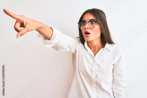 Young beautiful businesswoman wearing glasses standing over isolated white background Pointing with finger surprised ahead, open mouth amazed expression, something on the front