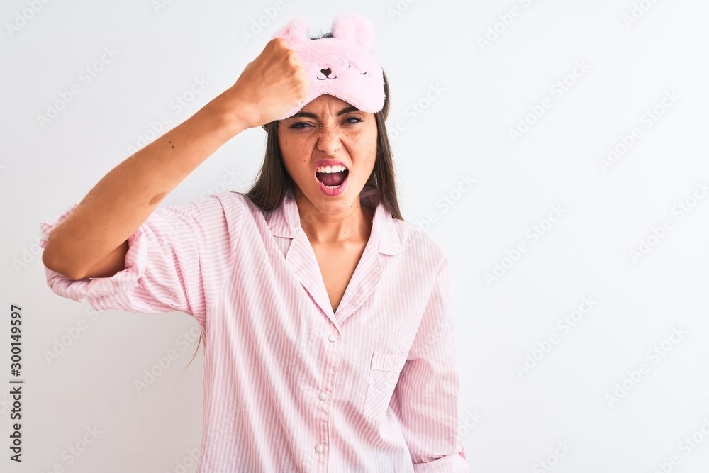 Young beautiful woman wearing sleep mask and pajama over isolated white background angry and mad raising fist frustrated and furious while shouting with anger. Rage and aggressive concept. Young beautiful woman wearing sleep mask and pajama over isolated white background angry and mad raising fist frustrated and furious while shouting with anger. Rage and aggressive concept.