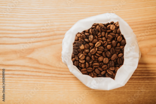 Roasted, brown coffee beans in white paper bag. Wooden background.