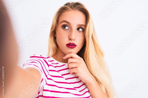 Beautiful woman wearing striped t-shirt make selfie by camera over isolated white background serious face thinking about question, very confused idea