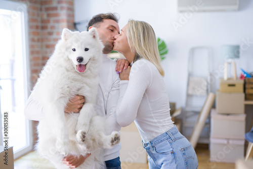 Fototapeta Naklejka Na Ścianę i Meble -  Young beautiful couple with dog kissing standing at new home around cardboard boxes