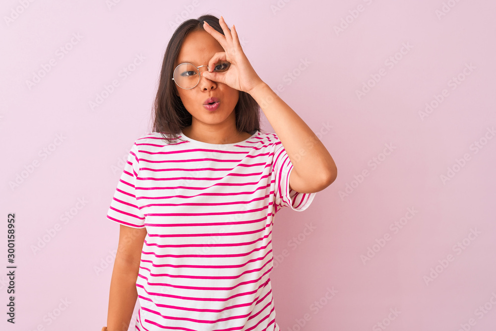 Young chinese woman wearing striped t-shirt and glasses over isolated pink background doing ok gesture shocked with surprised face, eye looking through fingers. Unbelieving expression. Young chinese woman wearing striped t-shirt and glasses over isolated pink background doing ok gesture shocked with surprised face, eye looking through fingers. Unbelieving expression.