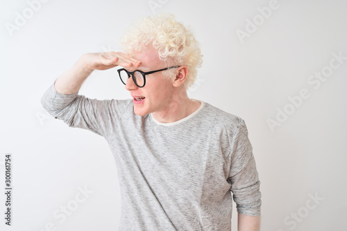 Young albino blond man wearing striped t-shirt and glasses over isolated white background very happy and smiling looking far away with hand over head. Searching concept.