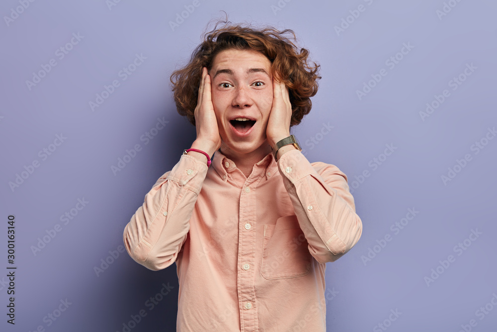 Overjoyed handsome man with long wavy hair, holding head with both hands, looking at camera with opened mouth, finds the solution of problem, expressing happiness, portrait, violet background Overjoyed handsome man with long wavy hair, holding head with both hands, looking at camera with opened mouth, finds the solution of problem, expressing happiness, portrait, violet background