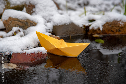 Yellow paper boat stuck in the ice, on a frozen puddle.
