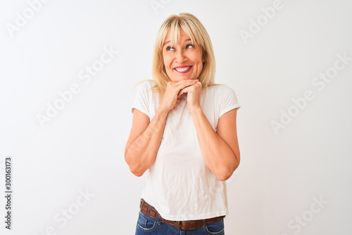 Middle age woman wearing casual t-shirt standing over isolated white background laughing nervous and excited with hands on chin looking to the side